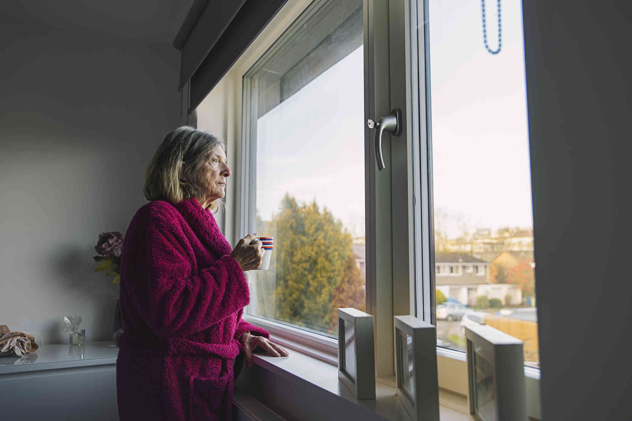 A mature woman in a red robe stands in her kitchen with a mug of coffe in her hands as she gazes sadly out of the window.